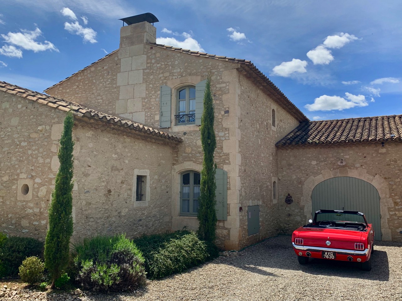 A red vintage convertible car parked outside a rustic stone house with arched garage doors, surrounded by clear blue skies and ornamental shrubs.